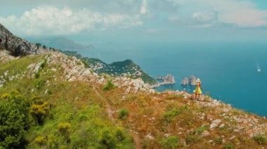 Bereketli yeşillik ve engebeli kayalar, mavi deniz manzaralı, tekneleri ve uzaktaki kayalık Faraglioni yığınları. İtalya, Capri 'de yürüyüş yapan bir kadın...