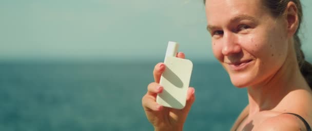 女人带着微笑开玩笑地推出防晒霜瓶 站在柔软的大海背景下 强调一种无忧无虑的 准备好海滩的态度 同时强调产品的重要性 — 图库视频影像