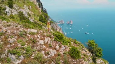 Capri Adası 'nın nefes kesici hava manzarası. Yeşil manzarada yürüyüş yapan bir kız. Denize ve dağınık teknelere bakıyor. Ünlü Faraglioni arka planda sallanıyor..