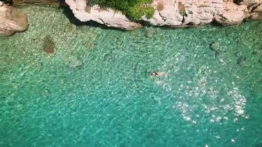Yalnız bir yüzücü kadın kayalık bir sahilin yakınındaki berrak suların tadını çıkarıyor. Durgun okyanusun havadan görünüşü..
