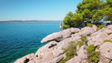 Kadın kayalık bir çıkıntıda duruyor ve engin denizi seyrediyor. Kıyı şeridi uzak mesafeye doğru kıvrılır, güneşli yaz gününde mavi gökyüzü..