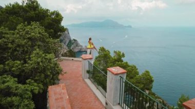 Sarı elbiseli bir kadın manzaralı bir manzaranın üstünde oturuyor. Kız, birden fazla teknenin yüzeyi işaretlediği sakin deniz manzarasına bakıyor. Capri İtalya..