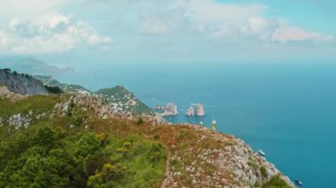 Capri, İtalya 'da Sahildeki Uçurumlara Bakan Yürüyüşçü Faraglioni. Kayaların üzerinde yürüyen bir kadın, kayıklarla benekli çarpıcı bir deniz manzarasına bakıyor...
