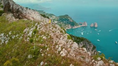 Yalnız bir figür, ikonik deniz yığınları Faraglioni olan Capri Adası 'nı görür. Dağlık araziye tünemiş, kıyı manzarası boyunca yürüyüş yapan bir kadın. İtalya 'da yaz tatilleri..