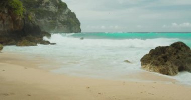 Köpüren dalgalar el değmemiş kumlu kıyı şeridini yıkıyor. Fırtınalı okyanustan deniz köpüğü geliyor. Uzak egzotik bir ada. Yavaş çekimde yakın çekim. Bali 'deki Tembeling plajı ve ormanı, Nusa Penida, Endonezya.