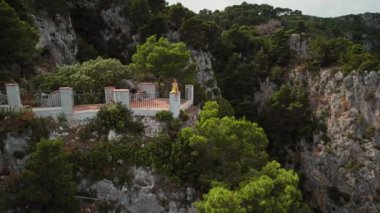 Uçurumun kenarındaki korkuluklara tünemiş bir insan, aşağıdaki gök mavisi okyanusu gözlemler. Yeşil ağaçlar ve sarp kayalıklar, panoramik deniz manzarasını çevreleyip geçen teknelerle donatır. Capri İtalya..