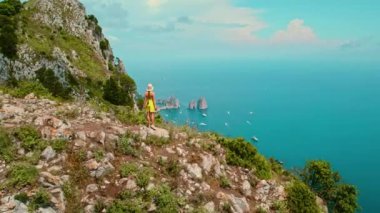 Turist uçurumun kenarında duruyor ve Capri Adası 'na bakıyor. Yeşil yemyeşil bitki örtüsü, kayalık kıyı ve çeşitli yelkenli gemilerle dağılmış huzurlu deniz...