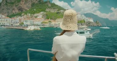 Amalfi şehrinin arka planında, teknede neşeli bir kadın. Güneş gözlüklü ve hasır şapkalı gülümseyen bir bayanın arkasında canlı bir kıyı şeridi ve masmavi sular...