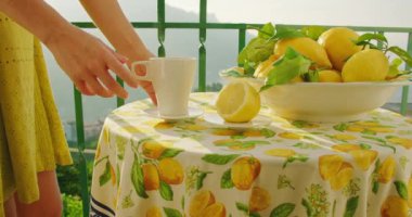 Bir insan eli, balkonda bir kase limonun yanında bardak tutar. Bu jest, güzel bir yerde huzurlu bir sabah geçirdiğimizi gösteriyor...