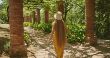 Tuğla sütunlarla desteklenen gölgeli bir pergola altında bir kadın neşeyle yürüyor, sakin bir bahçede yemyeşil bir bahçeyle çevrili...