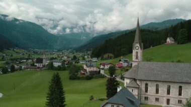 Önünde önemli bir kilise kulesi olan geniş bir vadi. Köy evleri ve bulutlu bir gökyüzünün altındaki orman tepeleriyle çevrili...