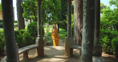 Çevresi yemyeşil ve yaz bahçesindeki tarihi kalıntılarla çevrili antik taş çardağı gezen bir kadın...