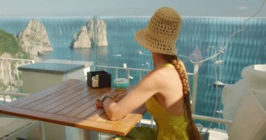 Şapkalı kadın, güneşli yaz gününde İtalya 'nın Capri Adası' ndaki terastan deniz manzarasının keyfini çıkarıyor..