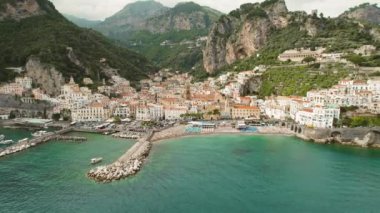 Amalfi kasabası ve limanı, renkli binalar, plaj şemsiyeleri ve dik yeşil uçurumlar ile deniz manzaralı bir zemin oluşturuyor...