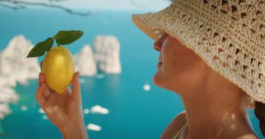 Hasır şapkalı kadın, İtalya 'ya yaz gezisi sırasında Faraglioni kayalıkları ve tekneleriyle Capri Adası' nın arka planındaki limona hayran...
