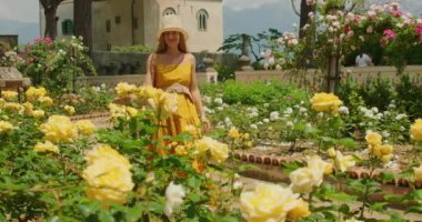 Sarı elbiseli ve hasır şapkalı bir kadın bahçedeki çiçeklerle dolu sarı gülleri koklamak için eğildi...