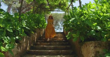 Yaz bahçesinin yemyeşil dalları arasında alçalan taş basamaklar. Sarı elbiseli ve güneş şapkalı kadın Ravello 'da yoğun yeşilliklerle çevrili bir şekilde yürür...