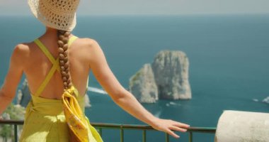 Elbise ve hasır şapkalı bir kadın, İtalya, Capri 'deki Faraglioni kaya oluşumlarına hayran, arkasında deniz olan korkuluklara yaslanmış...