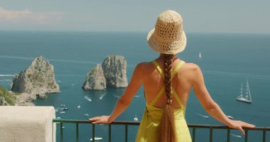 Şapkalı kadın Capri İtalya 'daki terastan Faraglioni kayalarına bakıyor. Turist yaz mevsiminin tadını çıkarıyor..