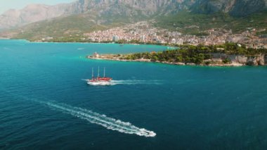 Hırvatistan 'ın Makarska kıyı kasabası yakınlarındaki kristal mavi deniz sularında ve yüksek dağ Biokovo' da bir kırmızı gemi yol alıyor. Yaz eğlence teknelerinin havadan görünüşü..