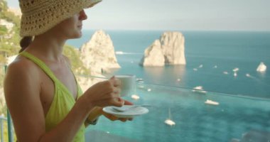 Şapkalı kadın, yaz güneşli bir günde İtalya 'nın Capri Adası' ndaki Faraglioni kayalıklarına bakan terasta kahvenin tadını çıkarıyor..