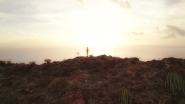 Turuncu elbiseli bir insanın silueti. Kolları havada okyanusun üzerinde gün doğumuna bakan kayalık bir arazide duruyor..