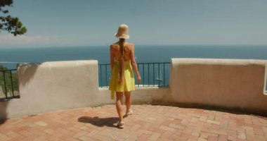 Uzun örgülü sarı elbiseli kadın denize bakan terasta yürüyor ve Faraglioni, Capri İtalya 'da sallanıyor...