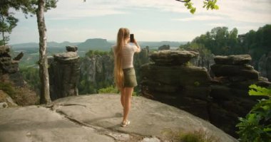Kadın telefonu ile panoramik bir görüntü yakalıyor. Bastei kaya oluşumları ve çevreleyen orman manzarası..
