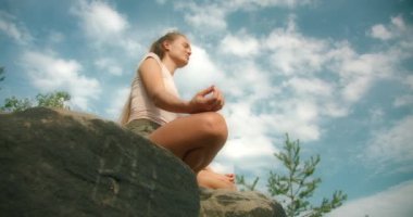 Bir kadın, bir nilüfer pozisyonunda, parlak bir gökyüzünün altındaki kayanın üzerinde, dağılmış bulutlarla, çam ağaçlarıyla çevrili bir şekilde meditasyon yapıyor...