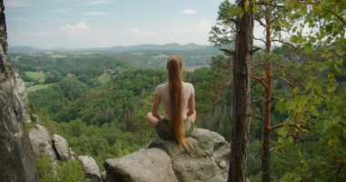 Uzun saçlı bir kadın bir kayanın üzerinde oturuyor, elleriyle bir kalp yapıyor, yemyeşil, geniş bir orman vadisine ve uzak dağlara bakıyor...