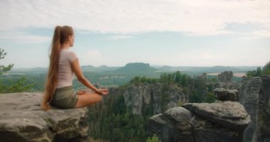 Uzun saçlı bir kadın, bir kayanın üzerinde lotus pozisyonunda oturur ve Bastei kaya oluşumlarının panoramik manzarasına ve engin orman manzarasına bakar...