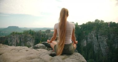 Uzun saçlı bir kadın, dramatik Bastei kaya oluşumlarına ve bereketli orman manzarasına göz yumarak lotus pozisyonunda meditasyon yapıyor...