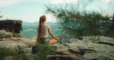 Bir kadın, Elbe Kum Taşı Dağları 'nın resimli manzarasıyla kayalık bir çıkıntıda meditasyon yapıyor. Uzun saçları sırtından aşağı süzülüyor...