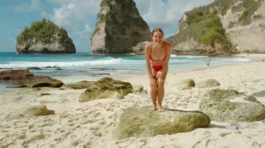 Pembe bikinili gülümseyen kadın kameraya el sallıyor ve deniz dalgalarına koşuyor, arkasında kayalıklar ve okyanuslar var. Nusa Penida Bali 'deki elmas plajı..