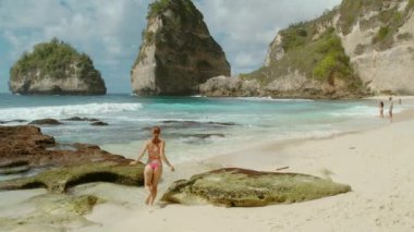 Pembe bikinili bir kadın yüksek uçurumlarla çevrili kristal berrak okyanusa doğru kumlu sahil boyunca yürüyor. Elmas plajı Nusa Penida Bali.