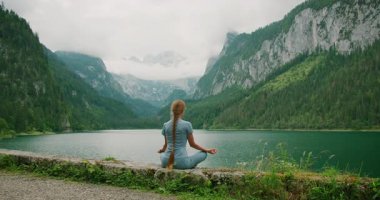 Çapraz bacaklarla meditasyon yapan bir kadın, Avusturya 'da yemyeşil ormanlarla çevrili bir dağ gölüne bakıyor..