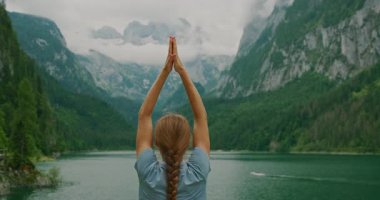 Huzurlu dağ gölü dramatik alp tepelerini yansıtıyor. Bir kadın kıyı kenarında yoga yapıyor...