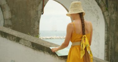 İtalya 'nın Amalfi kıyısındaki Atrani şehrindeki kemer köprüsünün altındaki merdivenlerden okyanusu gören bir kadın. Sarı elbiseli turist deniz kenarındaki haritayı inceliyor...