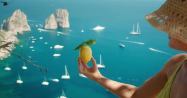 Hasır şapkalı kadın, İtalya 'nın Capri Adası' ndaki Faraglioni kaya oluşumları ve denizinde limon tutuyor..