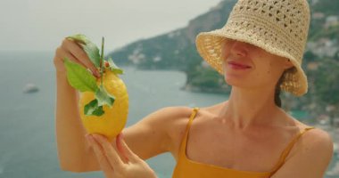 Arka planda deniz kayalıkları olan bir kadının tuttuğu limonun yakın çekimi..