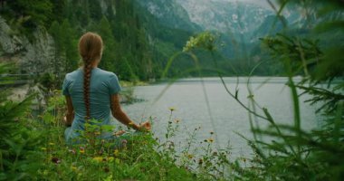 Vahşi çiçeklerin arasında oturmuş meditasyon yapan bir kadın, sakin bir alp gölü ve uzak dağlara bakıyor..