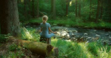 Huzurlu bir orman sahnesinde sakin bir derenin kenarında meditasyon yapan bir kadın.