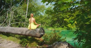 Hırvatistan 'ın Plitvice parkındaki turkuaz gölün üzerindeki kütüğün üzerinde meditasyonda oturan kadın.