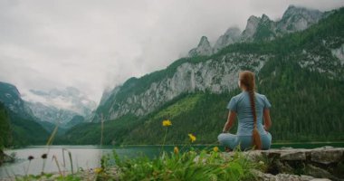 Kadın gölün kenarında oturur, sisli dağlarla çevrili, elleriyle kalp oluşturur..