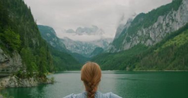 Saçı örgülü, göl kenarında oturan, manzaralı dağlara bakan bir kadının yakın çekimi. Dua eden kişi kollarını kaldırır...