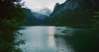 Bereketli yeşil ormanlarla çevrili sakin bir göl ve bulutlu bir gökyüzünün altında uzak dağlarla çevrili..