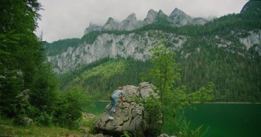 Orman ve dik dağ kayalıklarıyla çevrili canlı yeşil bir göl kenarındaki kayaya tırmanan bir insan..