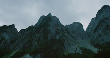 Karanlık dağ zirveleri bulutlu bir gökyüzünün altında, sık orman ve engebeli dağlık arazi ile çevrili..