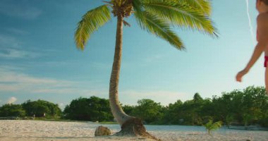 Tropikal güneş ışığında kumlu bir sahilde eğilmiş bir palmiye ağacına tırmanan bir kadın..