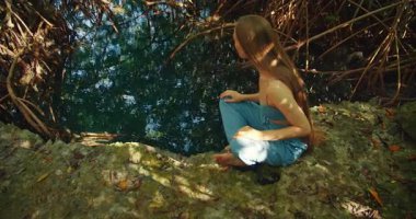 Kadın, sessiz bir mangrov gölünün kenarında yalınayak oturur ya da temiz suyla mezar taşı taşır..